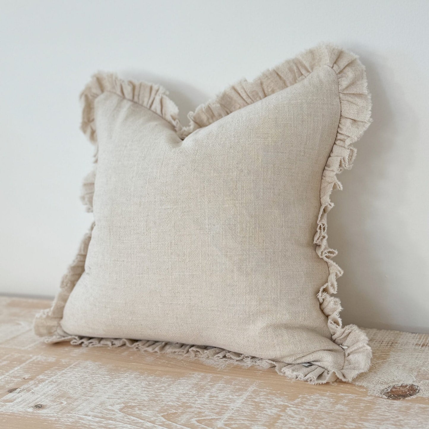 Beige pillow with ruffled edges on a wooden surface against a white wall