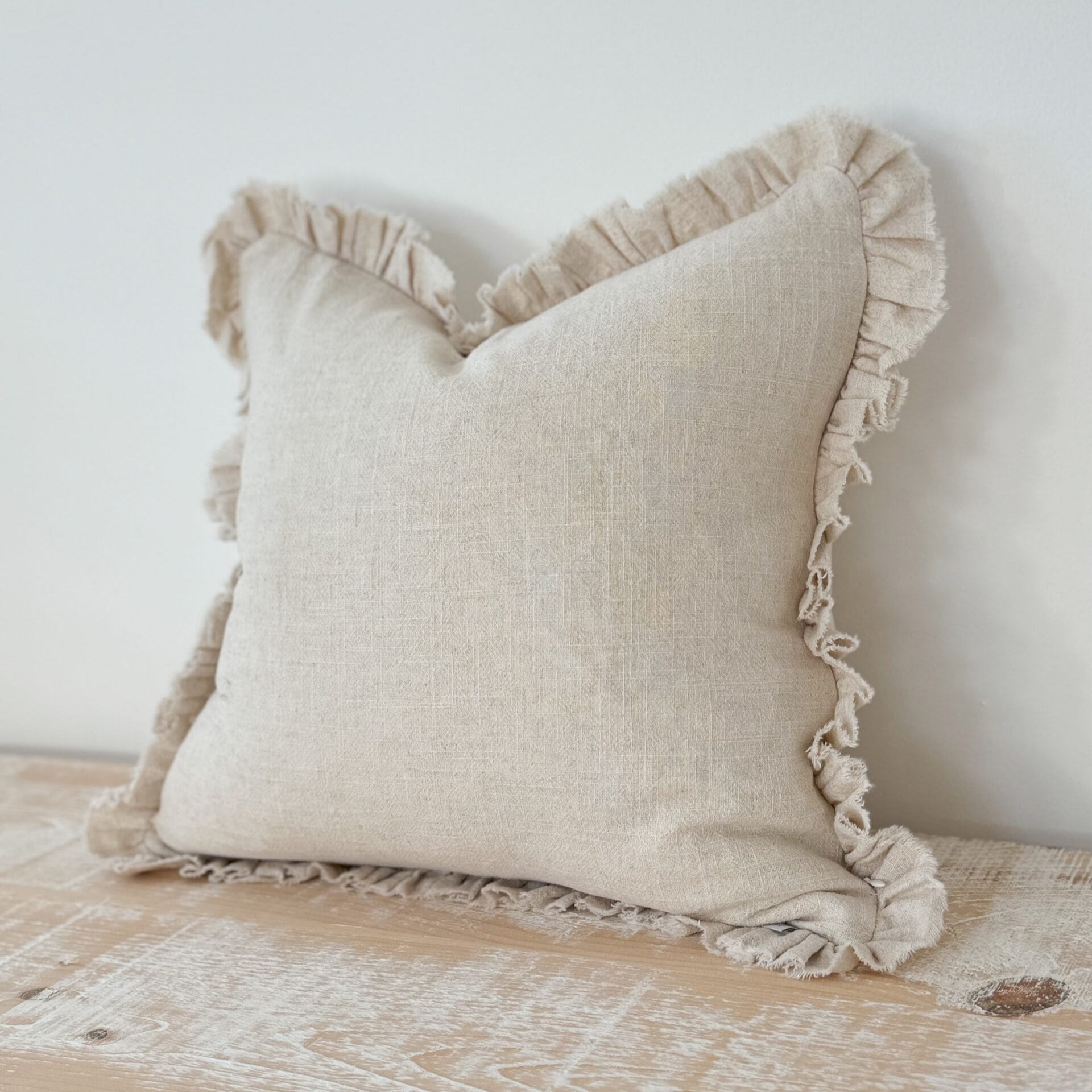 Beige pillow with ruffled edges on a wooden surface against a white wall